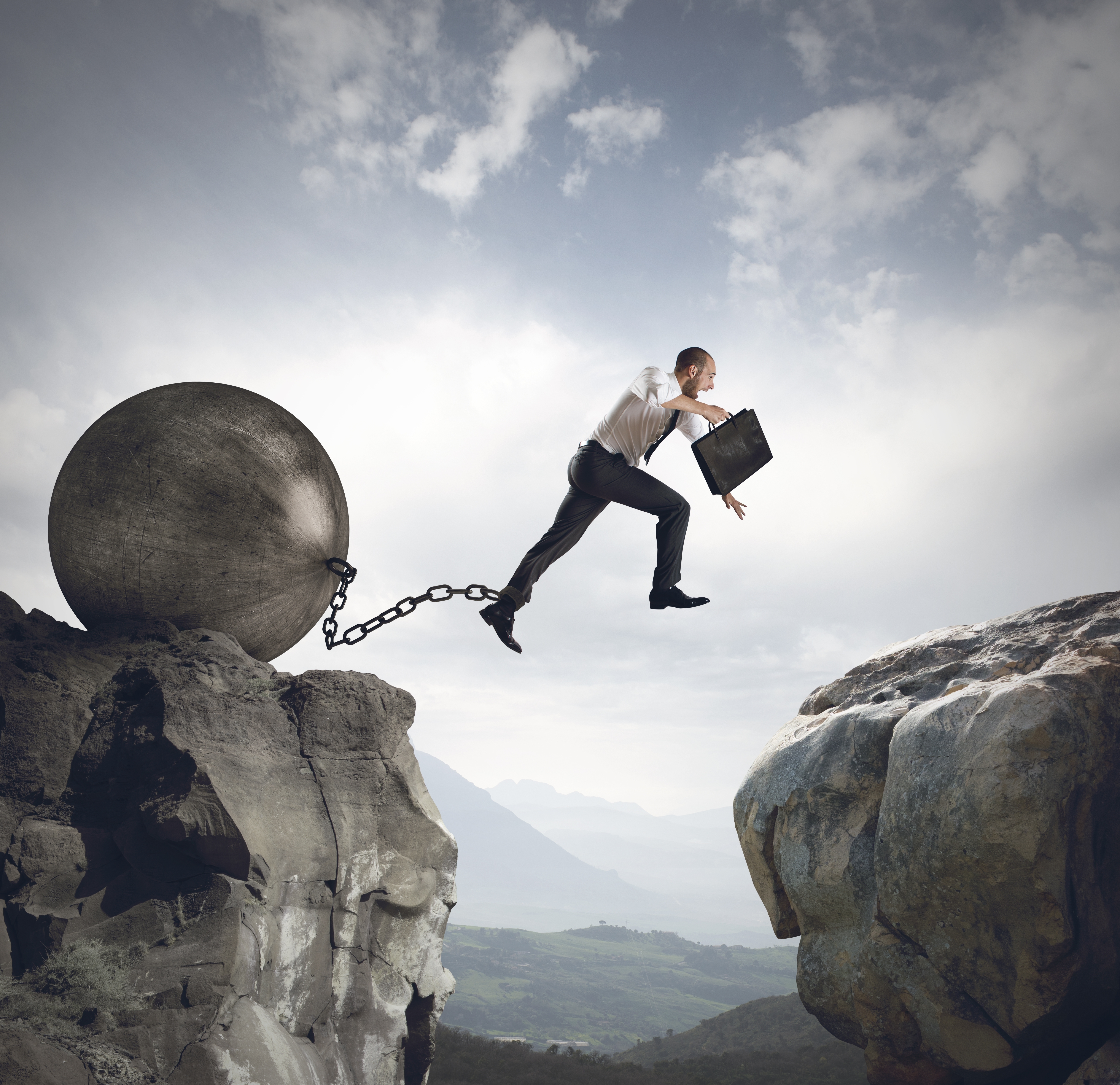 Business man with ball and chain attached to ankle jumping over large mountainous gap