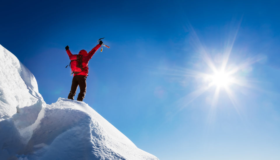 Person in snow gear with backpack standing on snowy peak raising arms in triumph.